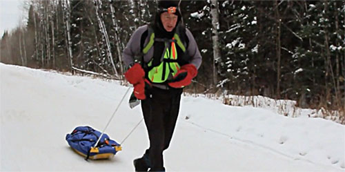 A man in yellow vest pulling a sled down the side of a snow covered slope.